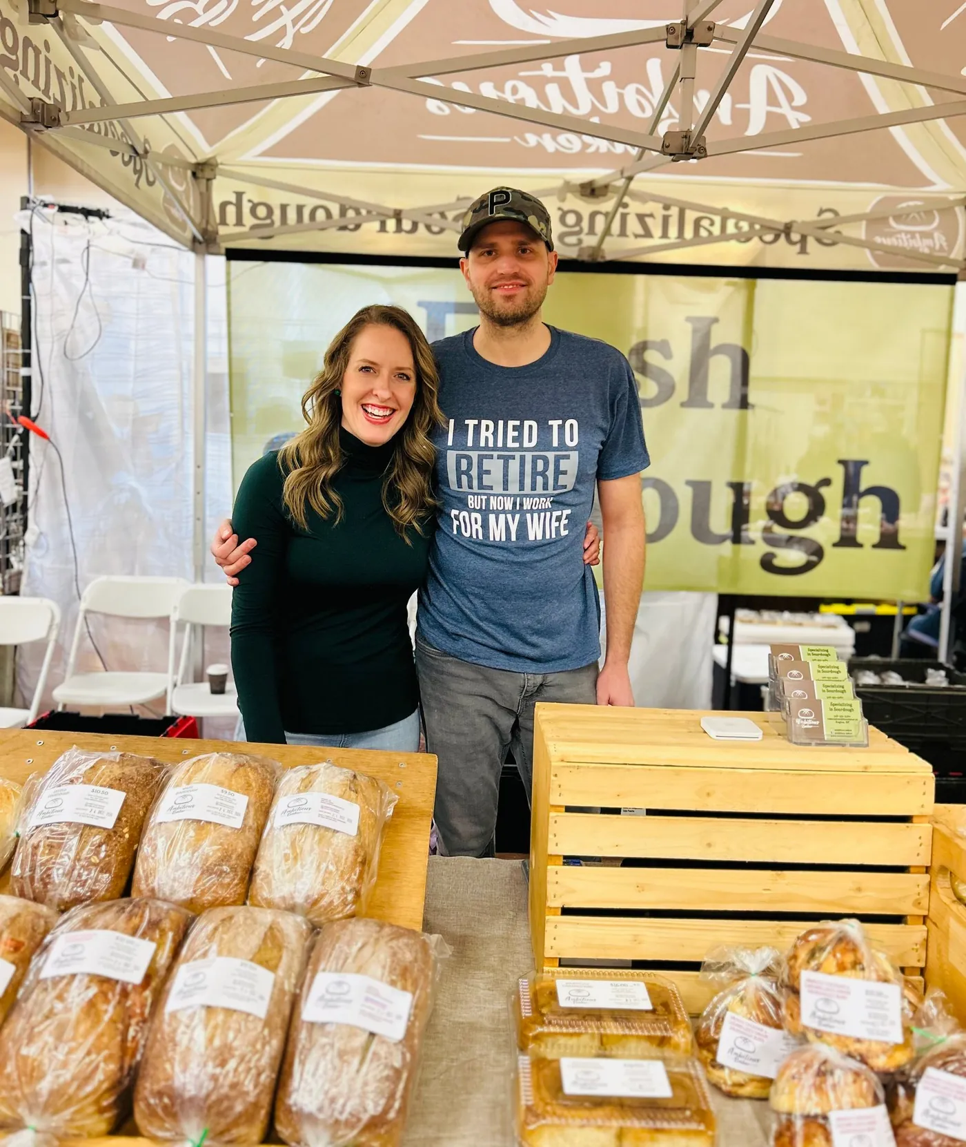 Quinn and Rebecca standing behind their market stall under the branded Ambitious Baker canopy, bagged loaves stacked on crates in front of them