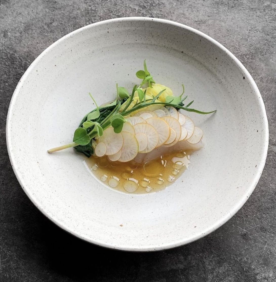 Delicate white-fish crudo with thin radish slices, microgreens and a clear citrus broth in a pale bowl, a refined first course, fine-dining photography for YQR Design
